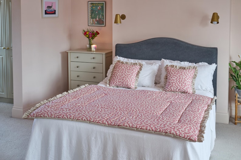 May include: A bed with a gray headboard, white bedding, and a red and white patterned quilt. Two matching throw pillows with ruffled edges sit on top. The room has pink walls and decorative wall art.