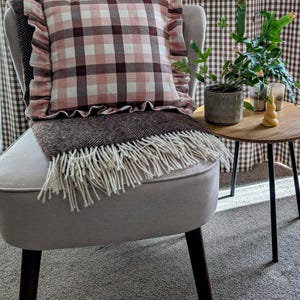 K&ouml;nnte beinhalten: Ein hellgrauer Sessel mit einem braunen und rosafarbenen karierten Kissen mit R&uuml;schen. Eine braune Decke mit wei&szlig;en Fransen liegt &uuml;ber dem Sessel. Ein kleiner Holztisch mit einer Topfpflanze und Kerzen steht neben dem Sessel.