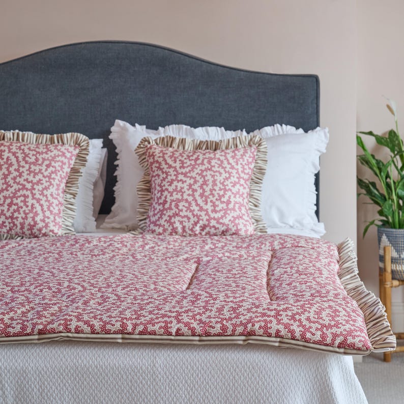 May include: A bed with a gray headboard, white bedding, and decorative pillows. The pillows and quilt feature a red and white floral pattern with ruffled edges. The quilt has a striped border. A potted plant is visible in the background.