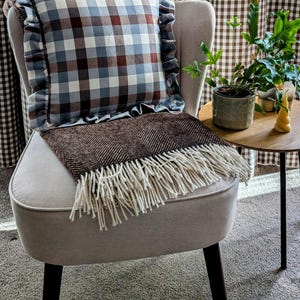 K&ouml;nnte beinhalten: Ein hellgrauer Sessel mit schwarzen Beinen, eine braune Wolldecke mit wei&szlig;en Fransen und ein blaues und braunes kariertes Kissen mit R&uuml;schen. Der Sessel steht vor einem braunen und wei&szlig;en Gingham-Vorhang.
