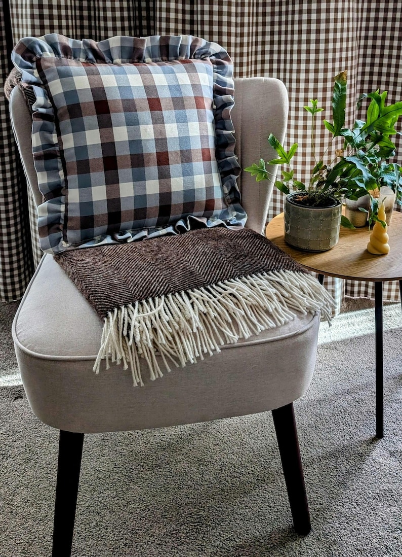 K&ouml;nnte beinhalten: Ein hellgrauer Sessel mit schwarzen Beinen, eine braune Wolldecke mit wei&szlig;en Fransen und ein blaues und braunes kariertes Kissen mit R&uuml;schen. Der Sessel steht vor einem braunen und wei&szlig;en Gingham-Vorhang.