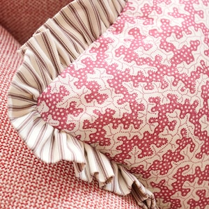 May include: Close-up of a decorative pillow with a red and white floral pattern. The pillow has a ruffled edge with a striped fabric. The pillow is resting on a red textured surface.