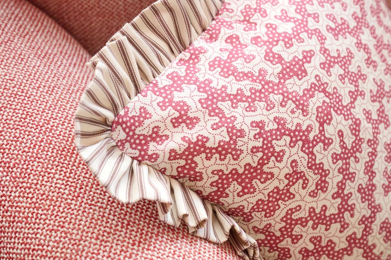 May include: Close-up of a decorative pillow with a red and white floral pattern. The pillow has a ruffled edge with a striped fabric. The pillow is resting on a red textured surface.