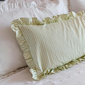 Könnte beinhalten: Ein rechteckiges Kissen mit grün-weißem Streifenmuster und Rüschenkanten. Das Kissen liegt auf einem weißen Bett, mit einem weiteren weißen Kissen mit Rüschenkanten im Hintergrund. Dies ist ein dekorativer Bettwäscheartikel.