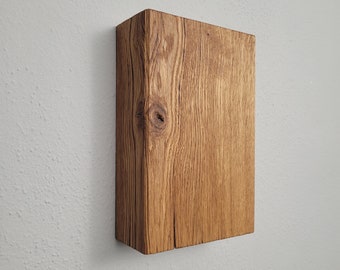 Puede incluir: Un bloque de madera rectangular y macizo con un acabado natural de color marrón claro. La veta de la madera es visible y el bloque está montado en una pared blanca. El bloque tiene un nudo y una grieta, lo que añade encanto rústico.