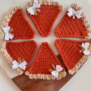 May include: Six crocheted pie slice coasters. Each slice is orange with a tan crust and a white and gold bow. The coasters are arranged on a wooden and marble surface. The theme is fall and Thanksgiving.