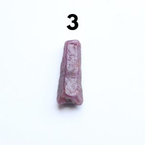 May include: A purplish-red gemstone, possibly a ruby, with a rough, elongated form, is set against a white backdrop. The number "3" is printed in black above the stone. The stone has a textured surface and an irregular shape.