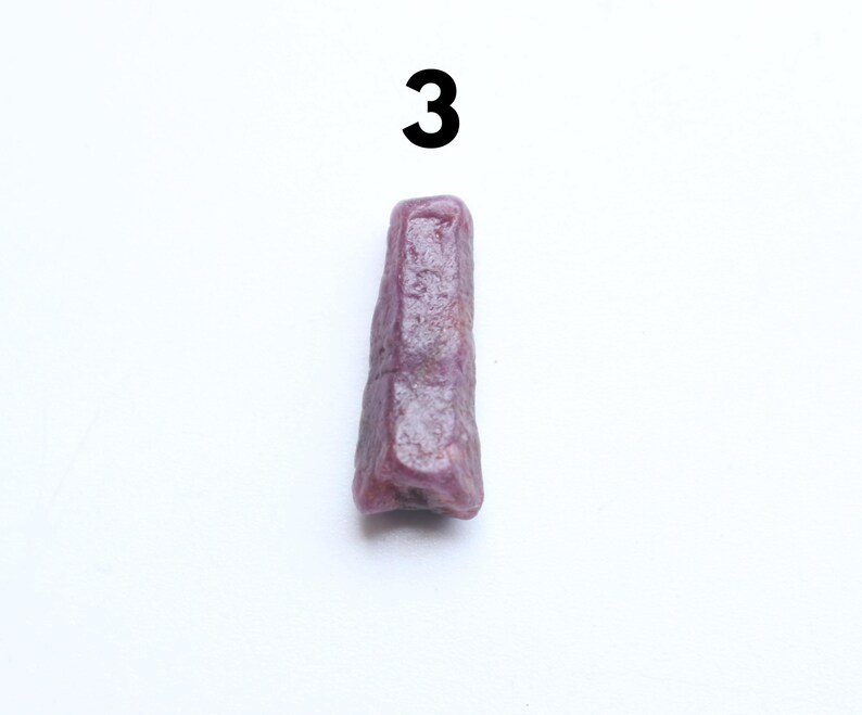 May include: A purplish-red gemstone, possibly a ruby, with a rough, elongated form, is set against a white backdrop. The number "3" is printed in black above the stone. The stone has a textured surface and an irregular shape.