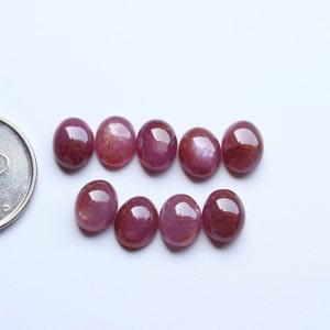 May include: Eight oval-shaped, pink ruby gemstones with a slight shimmer. The gemstones are arranged in two rows of four.