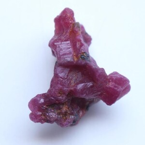 Könnte beinhalten: Ein roher, ungeschliffener Rubin-Edelstein mit tiefroter Farbe. Die natürliche Kristallformation hat eine raue, strukturierte Oberfläche und verschiedene Rot- und Brauntöne. Der Edelstein ist vor einem weißen Hintergrund platziert.