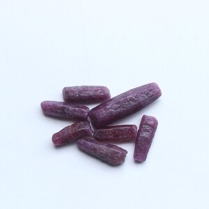 Pu&ograve; includere: Un gruppo di gemme di rubino grezze e non tagliate. Le gemme sono di un colore rosso-viola intenso e hanno una consistenza naturale ruvida. Le pietre sono di forma oblunga e variano leggermente in dimensioni, su sfondo bianco.