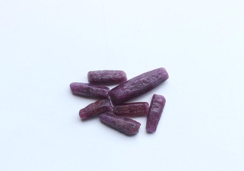 Pu&ograve; includere: Un gruppo di gemme di rubino grezze e non tagliate. Le gemme sono di un colore rosso-viola intenso e hanno una consistenza naturale ruvida. Le pietre sono di forma oblunga e variano leggermente in dimensioni, su sfondo bianco.