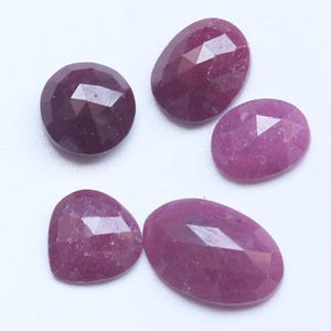 Puede incluir: Cinco piedras preciosas facetadas sueltas en varios tonos de magenta profundo y rosa oscuro. La colección incluye una piedra circular, tres piedras ovaladas y una piedra en forma de pera, todas con un acabado de superficie pulido y multifacetado.