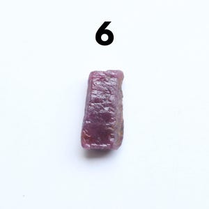 May include: A raw, uncut ruby gemstone with a deep reddish-purple hue. The rough stone has a textured surface and is displayed against a white background. The number "6" is above the gemstone.