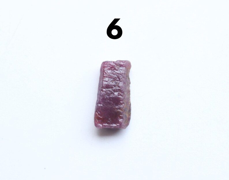 May include: A raw, uncut ruby gemstone with a deep reddish-purple hue. The rough stone has a textured surface and is displayed against a white background. The number "6" is above the gemstone.