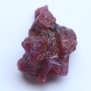 May include: A rough, uncut ruby gemstone with a deep red hue. The natural stone has an irregular, textured surface with visible crystalline structures. The ruby is set against a white background, highlighting its color and form.