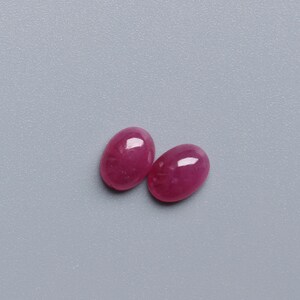 May include: Two oval-shaped, polished ruby gemstones. The rubies are a deep, vibrant red colour with a glossy finish, reflecting light. They are set against a plain, light grey background. These gemstones are suitable for jewellery making.