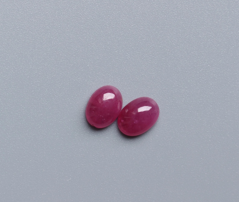 May include: Two oval-shaped, polished ruby gemstones. The rubies are a deep, vibrant red colour with a glossy finish, reflecting light. They are set against a plain, light grey background. These gemstones are suitable for jewellery making.
