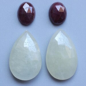 May include: Two oval, faceted, dark red gemstones are positioned above two pear-shaped, light green gemstones. The gemstones are displayed on a light grey surface. The gems are likely for jewellery making.