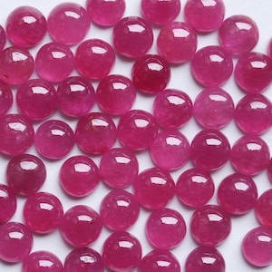Pode incluir: Uma coleção de pedras preciosas de rubi redondas e polidas. As gemas são de uma cor vermelho-rosada profunda, com uma superfície lisa e refletora. As pedras preciosas estão espalhadas pela imagem, criando uma exibição visualmente atraente.
