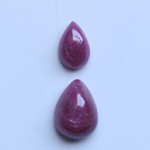 以下が含まれることがあります： 2つの研磨された、涙の形をした宝石。宝石は、光沢のある仕上げの深い、豊かな赤紫の色をしています。石は明るい背景に対して表示されています。