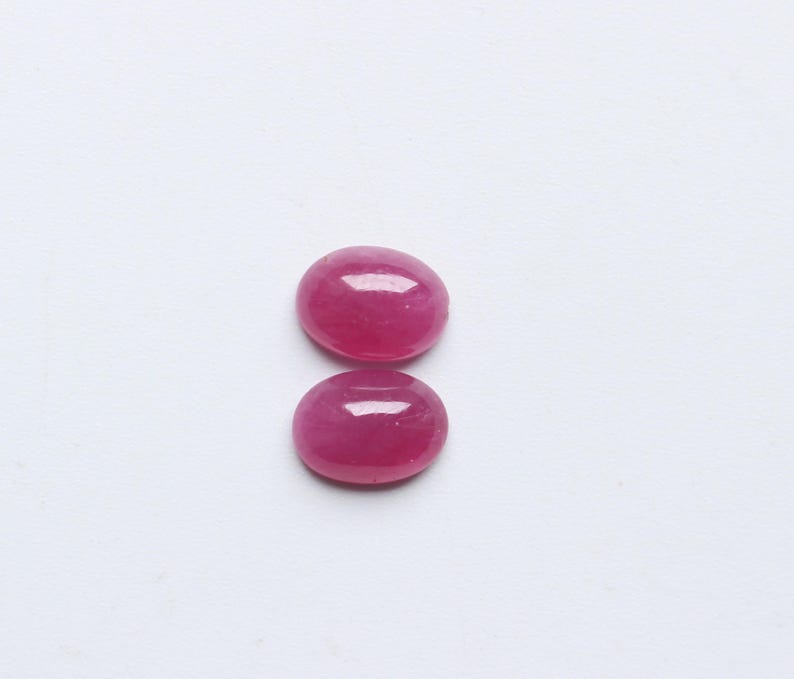 Op de afbeelding: Twee ovaalvormige, gepolijste robijn edelstenen. De edelstenen hebben een levendige, diepe roze-rode kleur, met een glad, glanzend oppervlak. Ze zijn geplaatst tegen een effen witte achtergrond, wat hun kleur en vorm benadrukt.