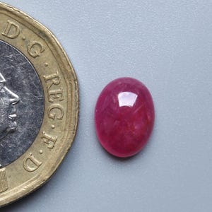 May include: A polished, oval-shaped, red gemstone next to a gold-coloured coin. The gemstone has a smooth surface and a vibrant colour. The coin features a profile of a person and the text "ELIZABETH II D.G. REG. F.D. 2017".