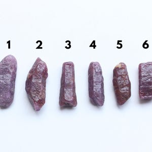 May include: Six raw ruby gemstones of differing shapes and sizes are displayed on a white surface. The gemstones vary in colour from deep purplish-red to lighter shades, with visible textures and natural formations. Numbers 1 to 6 are above each stone.