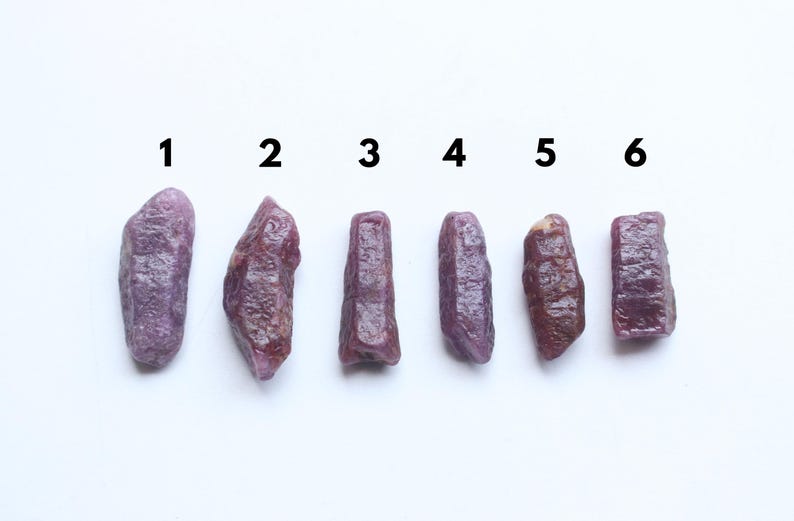 May include: Six raw ruby gemstones of differing shapes and sizes are displayed on a white surface. The gemstones vary in colour from deep purplish-red to lighter shades, with visible textures and natural formations. Numbers 1 to 6 are above each stone.