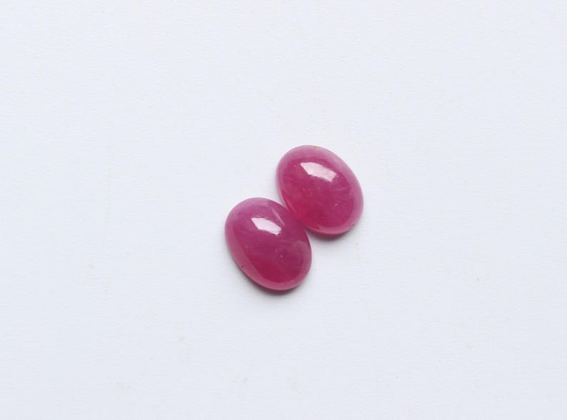 May include: Two oval-shaped, polished gemstones in a vibrant pink-red hue. The gems have a smooth, glossy surface, reflecting light. They are set against a plain white background, highlighting their colour and shape. These could be rubies or similar gemstones.