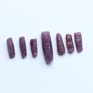 Pu&ograve; includere: Sette gemme di rubino grezze, non tagliate, di varie dimensioni e tonalit&agrave; rosso-viola scuro sono disposte su una superficie bianca. Le gemme hanno un aspetto naturale e strutturato, che mostra il loro stato grezzo.