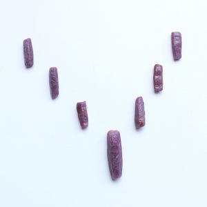 Pu&ograve; includere: Una collezione di otto gemme di rubino grezze, allungate. Le gemme sono di un colore rosso-viola intenso e sono disposte su uno sfondo bianco. Ogni pietra ha una superficie leggermente irregolare e strutturata.