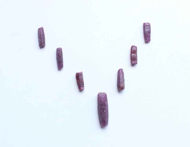 Pu&ograve; includere: Una collezione di otto gemme di rubino grezze, allungate. Le gemme sono di un colore rosso-viola intenso e sono disposte su uno sfondo bianco. Ogni pietra ha una superficie leggermente irregolare e strutturata.