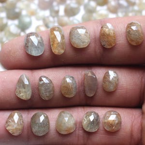 May include: A hand holds a selection of faceted gemstones in different shapes and sizes. The gems vary in colour from pale yellow to light brown, with some showing a hint of blue. The gemstones are displayed against a blurred background of similar stones.