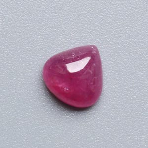 May include: A polished, cabochon-cut ruby gemstone. The gem is a deep, saturated pinkish-red color with a smooth, rounded shape. The surface reflects light, creating highlights. The gemstone is set against a neutral gray background.