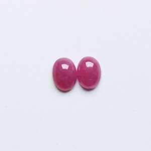 May include: Two oval-shaped, polished gemstones in a vibrant, deep pink hue. The gems are set against a plain white background, highlighting their smooth, reflective surfaces. These gemstones are likely for jewelry making.
