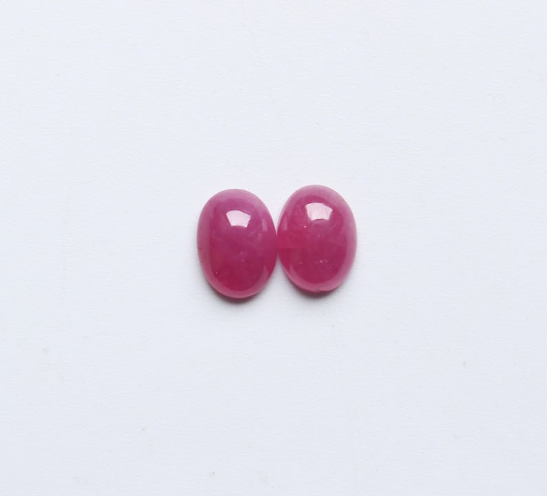 May include: Two oval-shaped, polished gemstones in a vibrant, deep pink hue. The gems are set against a plain white background, highlighting their smooth, reflective surfaces. These gemstones are likely for jewelry making.