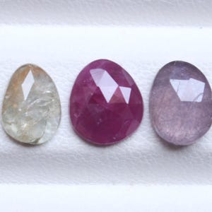 May include: Three faceted gemstones in a row. The first is a light brown and clear color, the second is a deep red, and the third is a light purple. The gemstones are oval-shaped and sit on a white surface.