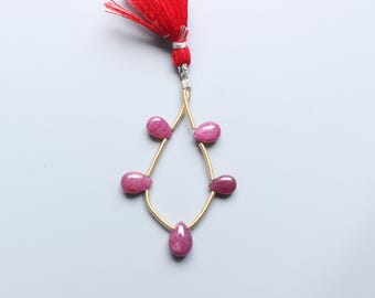 May include: A strand of pink, teardrop-shaped gemstones on a gold wire, with a red tassel. The gemstones are arranged in a teardrop shape. The background is a light grey colour.