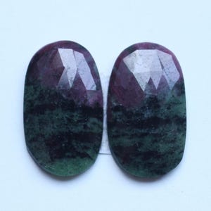 May include: Two oval-shaped gemstones with a faceted top. The stones are a mix of deep red and green hues, with a dark, mottled pattern. The gemstones are set against a white background.