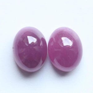 May include: Two oval-shaped, cabochon-cut gemstones with a smooth, polished surface and a vibrant, deep pinkish-purple hue. Each stone features a translucent quality with subtle internal light reflections, showcasing a rounded, dome-like profile.