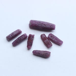 Pu&ograve; includere: Una collezione di gemme di rubino grezze e non tagliate. Le gemme sono di un colore rosso-viola intenso e ricco, con una superficie leggermente ruvida e strutturata. Le pietre sono di forma oblunga.