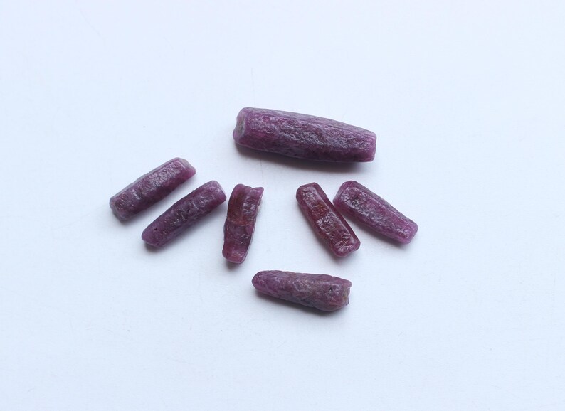 Pu&ograve; includere: Una collezione di gemme di rubino grezze e non tagliate. Le gemme sono di un colore rosso-viola intenso e ricco, con una superficie leggermente ruvida e strutturata. Le pietre sono di forma oblunga.