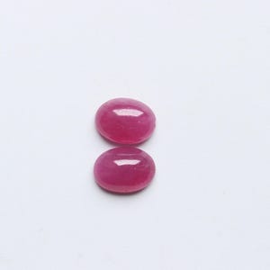 May include: Two oval-shaped, polished ruby gemstones. The rubies are a vibrant, deep pinkish-red colour, with a smooth, glossy surface. They are set against a plain white background, highlighting their colour and shape.