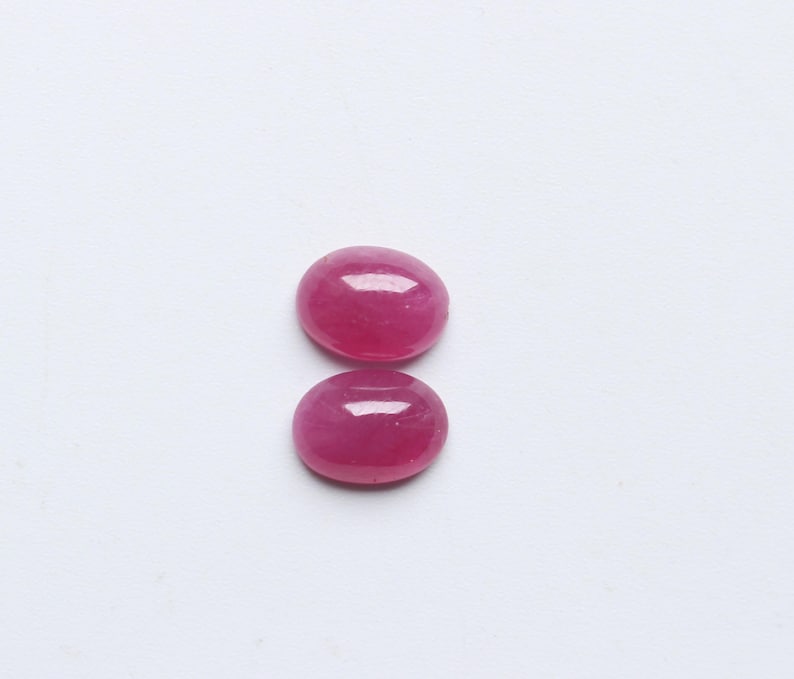 May include: Two oval-shaped, polished ruby gemstones. The rubies are a vibrant, deep pinkish-red colour, with a smooth, glossy surface. They are set against a plain white background, highlighting their colour and shape.