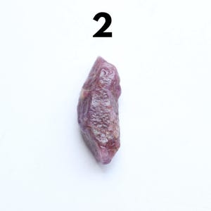 May include: A natural ruby gemstone, rough in form, with a deep purple colour. The ruby is elongated and has a textured surface. The number "2" is printed in black at the top of the image.