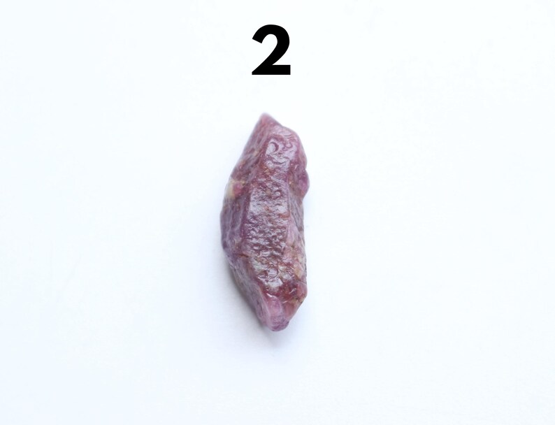 May include: A natural ruby gemstone, rough in form, with a deep purple colour. The ruby is elongated and has a textured surface. The number "2" is printed in black at the top of the image.