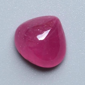 Puede incluir: Una piedra preciosa de rubí pulida, de forma ovalada. La gema es de un color rojo-rosado profundo y saturado, con una superficie lisa y reflectante. La piedra está sobre un fondo gris claro.