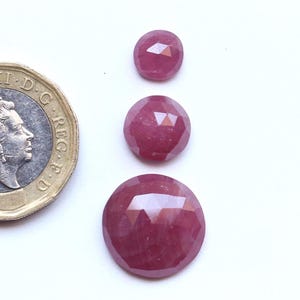 May include: Three round, faceted, deep pink gemstones arranged vertically by size, next to a British two-pound coin for scale. The stones exhibit a polished, multi-faceted surface that reflects light, showing a consistent dark magenta hue across all three pieces.