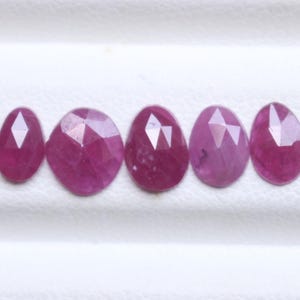 Puede incluir: Cinco gemas de rubí facetadas en varias formas ovaladas y redondas. Las gemas son de un color rosa-rojo intenso, con múltiples facetas que capturan la luz. Las gemas se muestran sobre una superficie blanca.